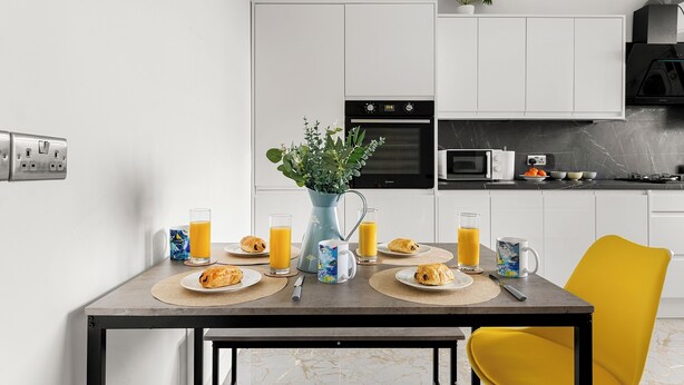 A modern kitchen with white cabinets, a black oven, and a microwave. There is a dining table with plates of food, glasses of orange juice, and a pitcher of water. - Hartley Avenue (Leeds)