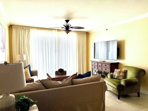 Una sala de estar con ventilador de techo, un sofá, un sillón verde, un televisor y una ventana con cortinas.