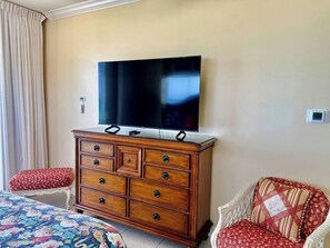 A hotel room with a flat-screen TV mounted on a wooden dresser, a bed with a floral bedspread, and a red patterned chair. - Sand-Toes Living | Gulf-Front, Walk to Pier Park (Panama City Beach)