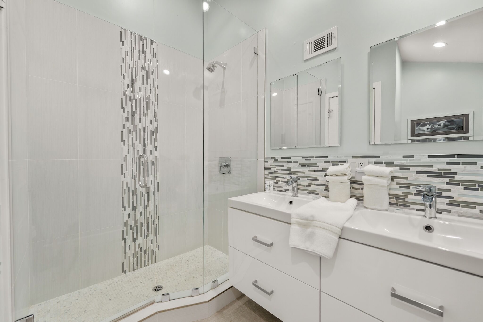 A modern bathroom with a glass shower enclosure, white vanity with two sinks, and a tiled backsplash.