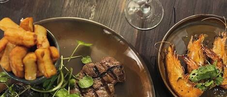 A dining table with a plate of steak, fries, and shrimp, accompanied by wine glasses and a lamp.