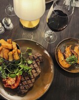 Una mesa de comedor con un plato de bistec, papas fritas y camarones, junto con copas de vino y una lámpara.