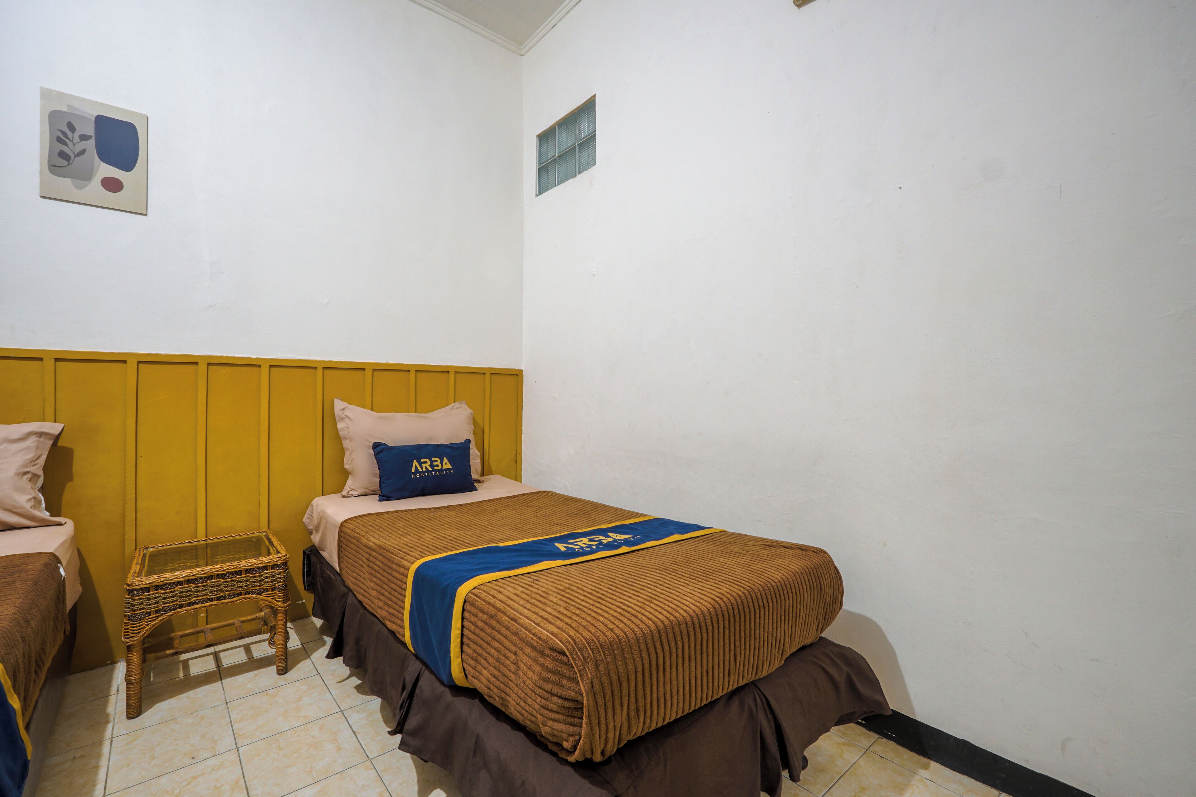 A small, simple room with two beds, a small table, and a wall-mounted shelf.