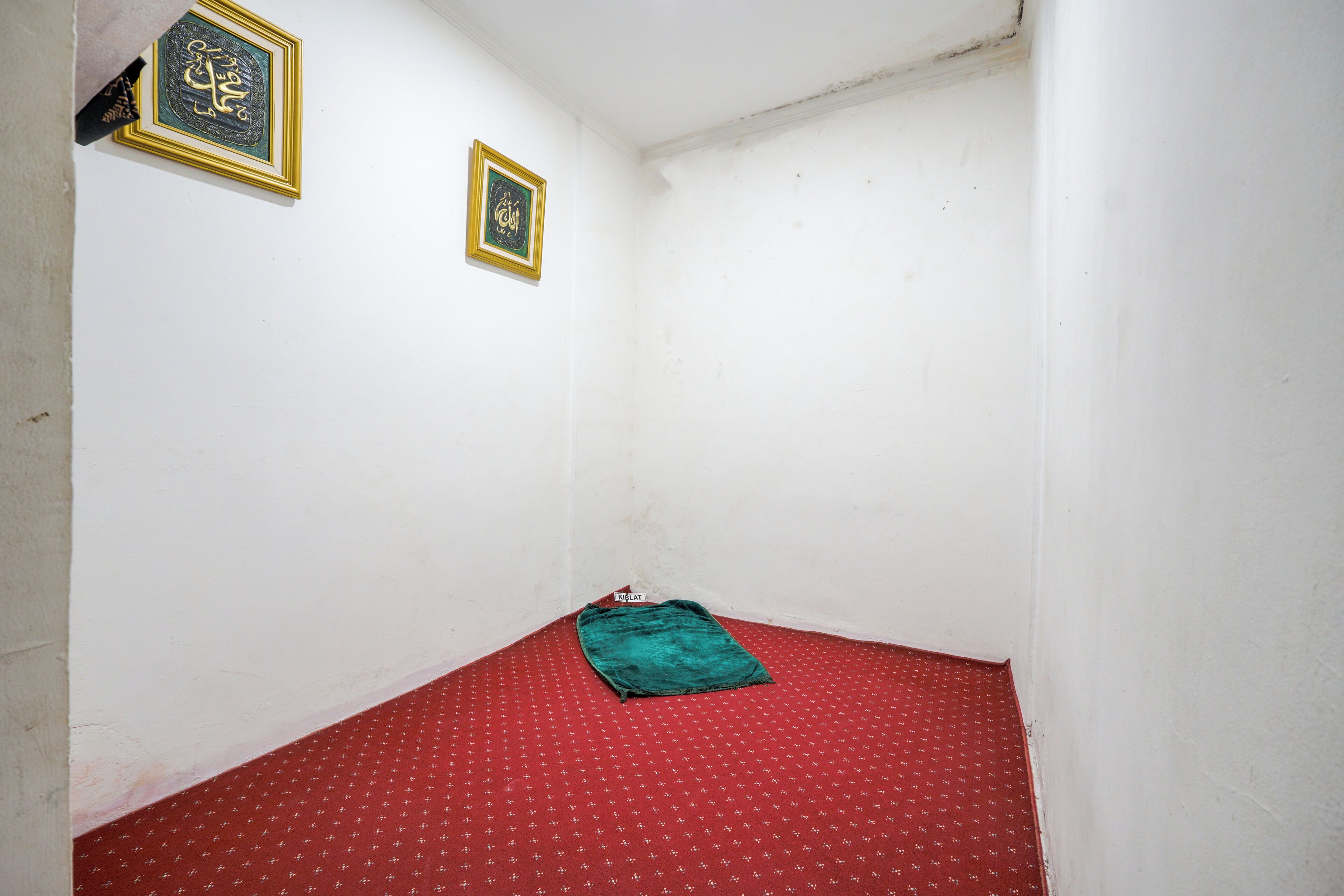 A small room with a red patterned carpet, two framed calligraphy pieces on the wall, and a green blanket on the floor.