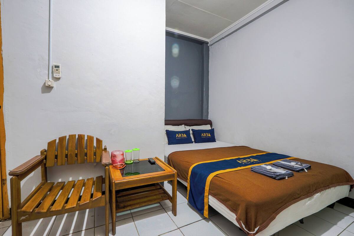 A small hotel room with a wooden chair, a small table, a bed with a brown bedspread, and a window with blinds.