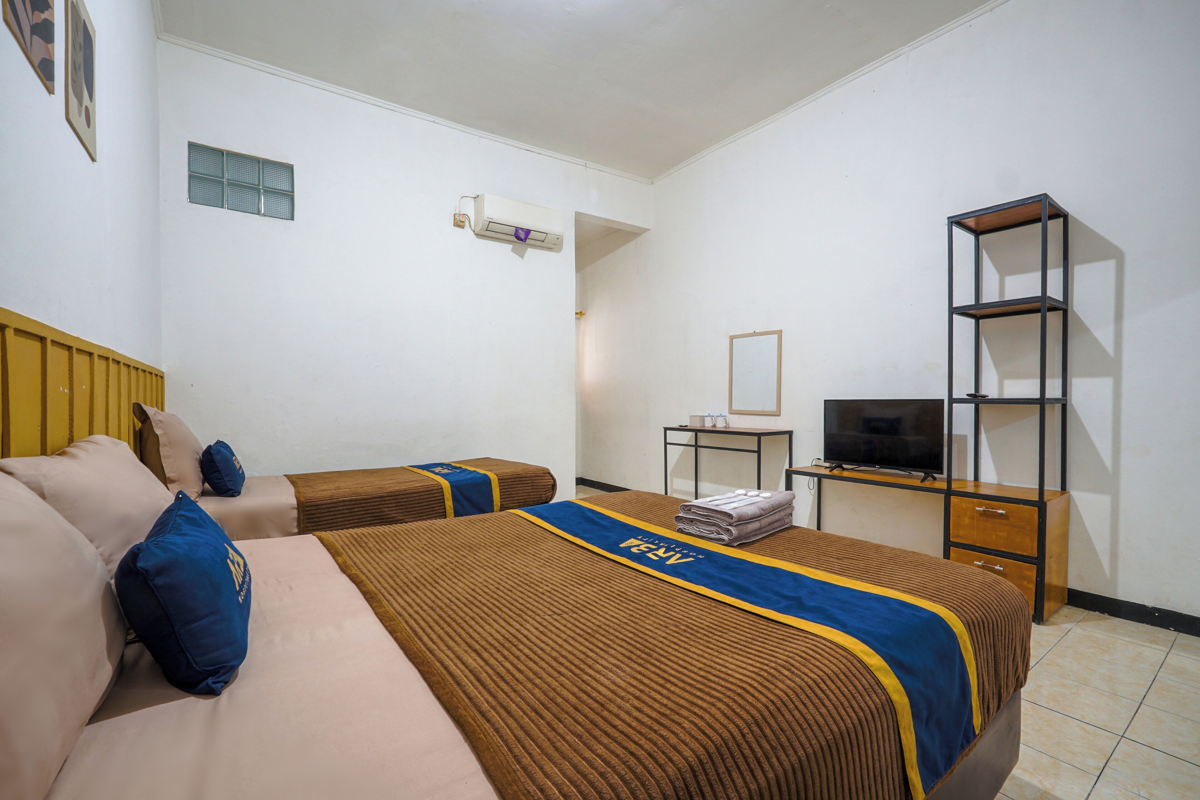 A hotel room with two beds, a TV, a small table, and a shelf.