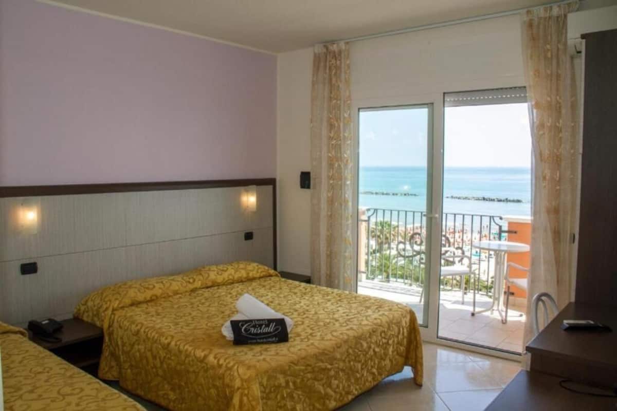 Habitación de hotel con dos camas, un balcón con vista al mar y una pequeña mesa.