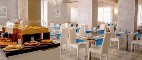 A hotel dining area with multiple tables and chairs, a buffet with various food items, and decorative chandeliers.