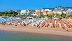 Una playa con hileras de sombrillas y sillas de descanso, edificios al fondo y agua cristalina azul.