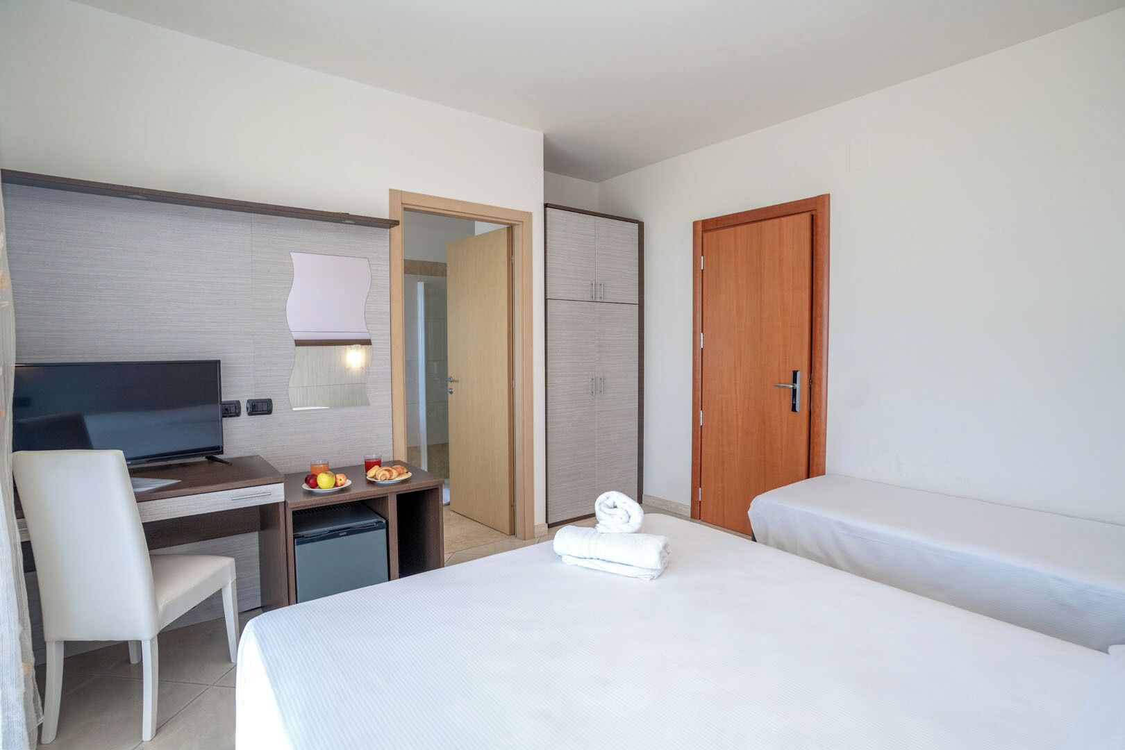 A hotel room with a bed, a desk with a TV, a chair, a small fridge, and a wooden door.