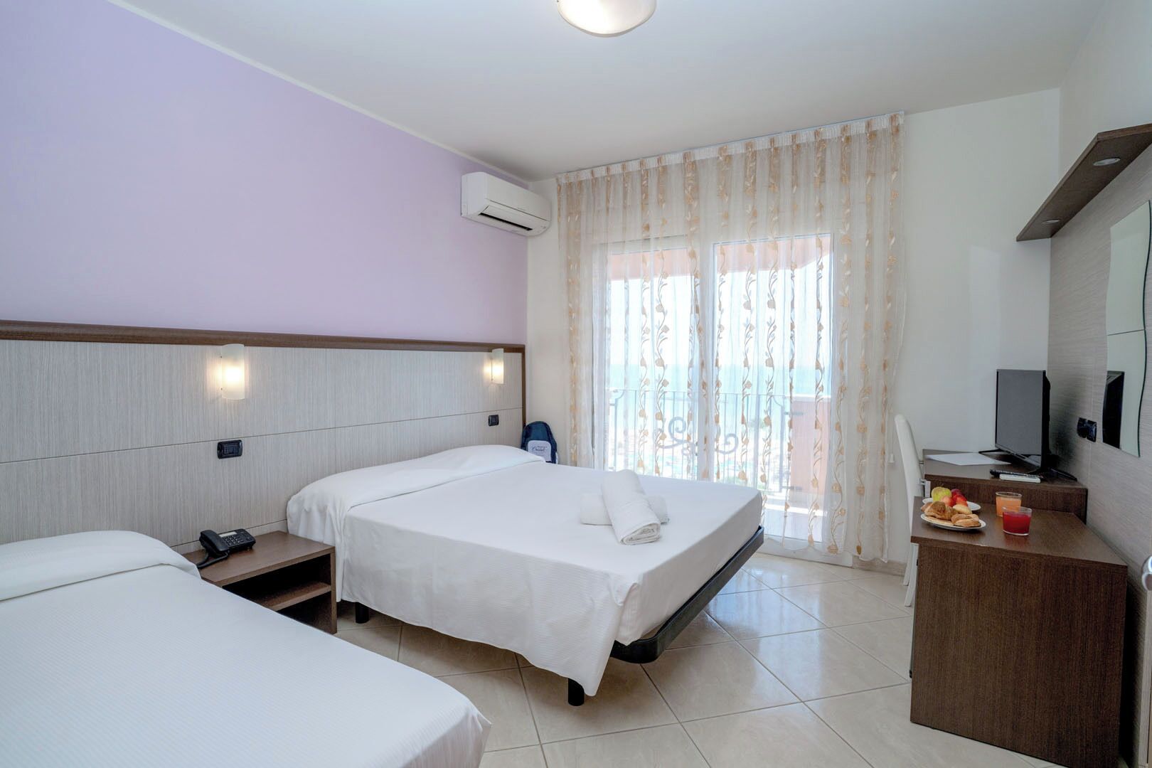A hotel room with two beds, a desk with a chair, a TV, and a balcony with curtains.