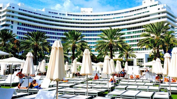 A modern hotel with a curved facade, outdoor swimming pool area with sun loungers and umbrellas, and palm trees. - Fontainebleau Jr. Suite Tresor just Renovated (Miami Beach)