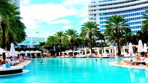 A large outdoor swimming pool surrounded by palm trees and lounge chairs, with a modern hotel building in the background. - Fontainebleau Jr. Suite Tresor just Renovated (Miami Beach)
