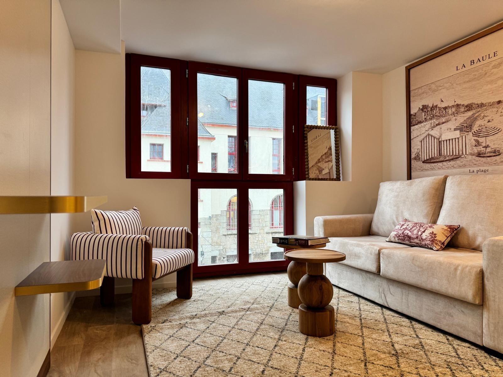 A living room with a striped sofa, a beige sofa, and a wooden coffee table. Large windows with red frames provide a view of buildings outside.