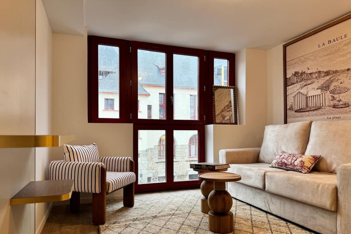 A living room with a striped sofa, a beige sofa, and a wooden coffee table. Large windows with red frames provide a view of buildings outside.
