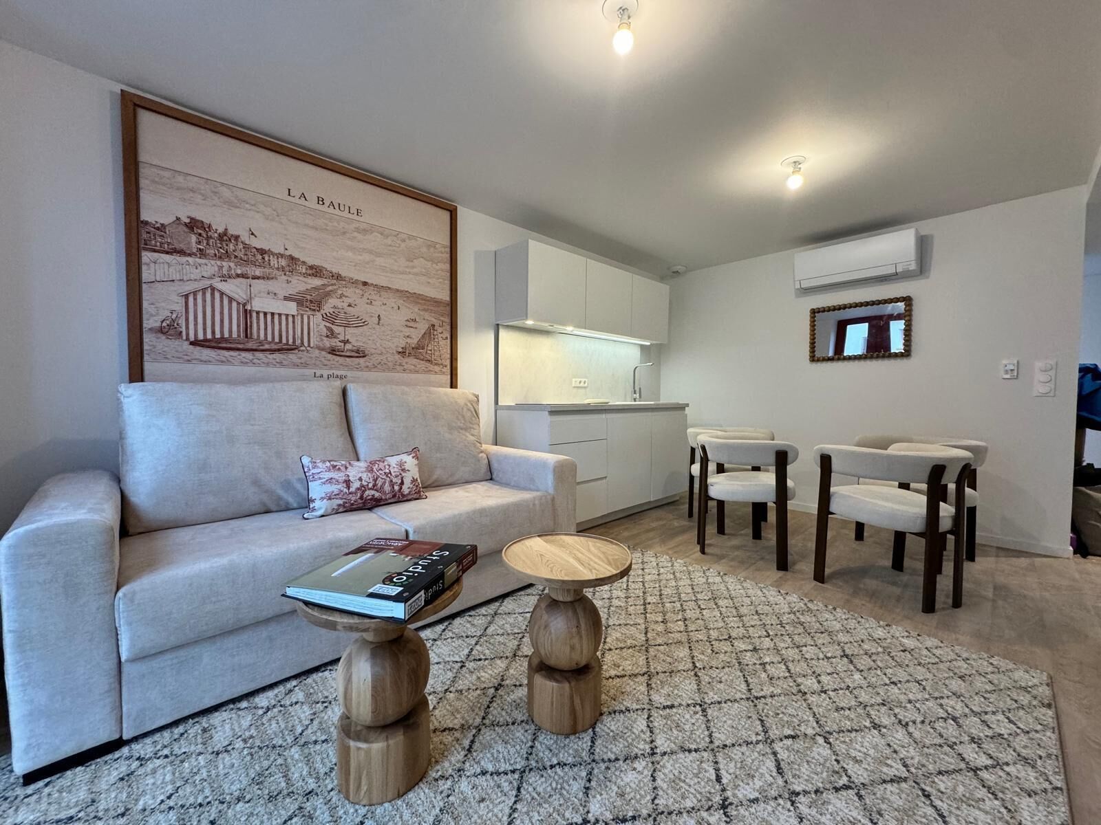 A modern living room with a sofa, a coffee table, and two wooden side tables. There is a kitchen area with white cabinets and a dining set. A framed picture of a beach scene is on the wall.