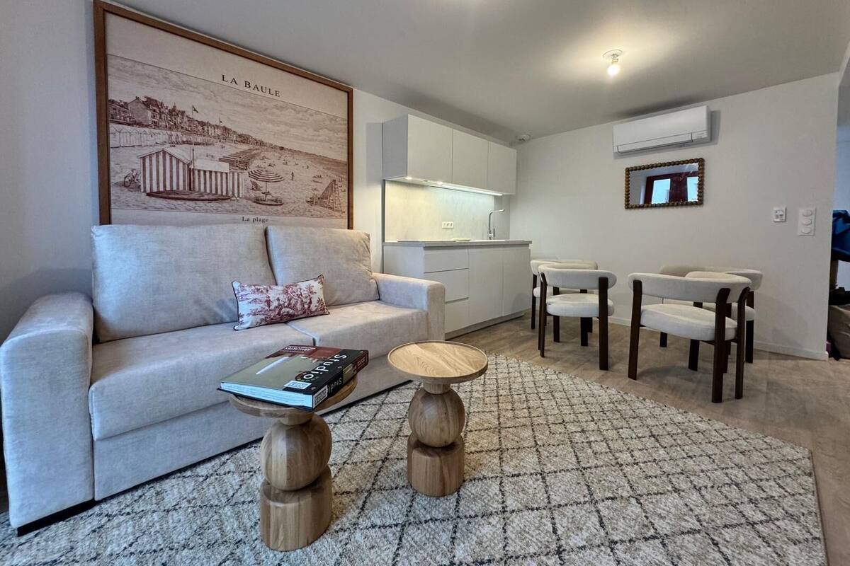 A modern living room with a sofa, a coffee table, and two wooden side tables. There is a kitchen area with white cabinets and a dining set. A framed picture of a beach scene is on the wall.