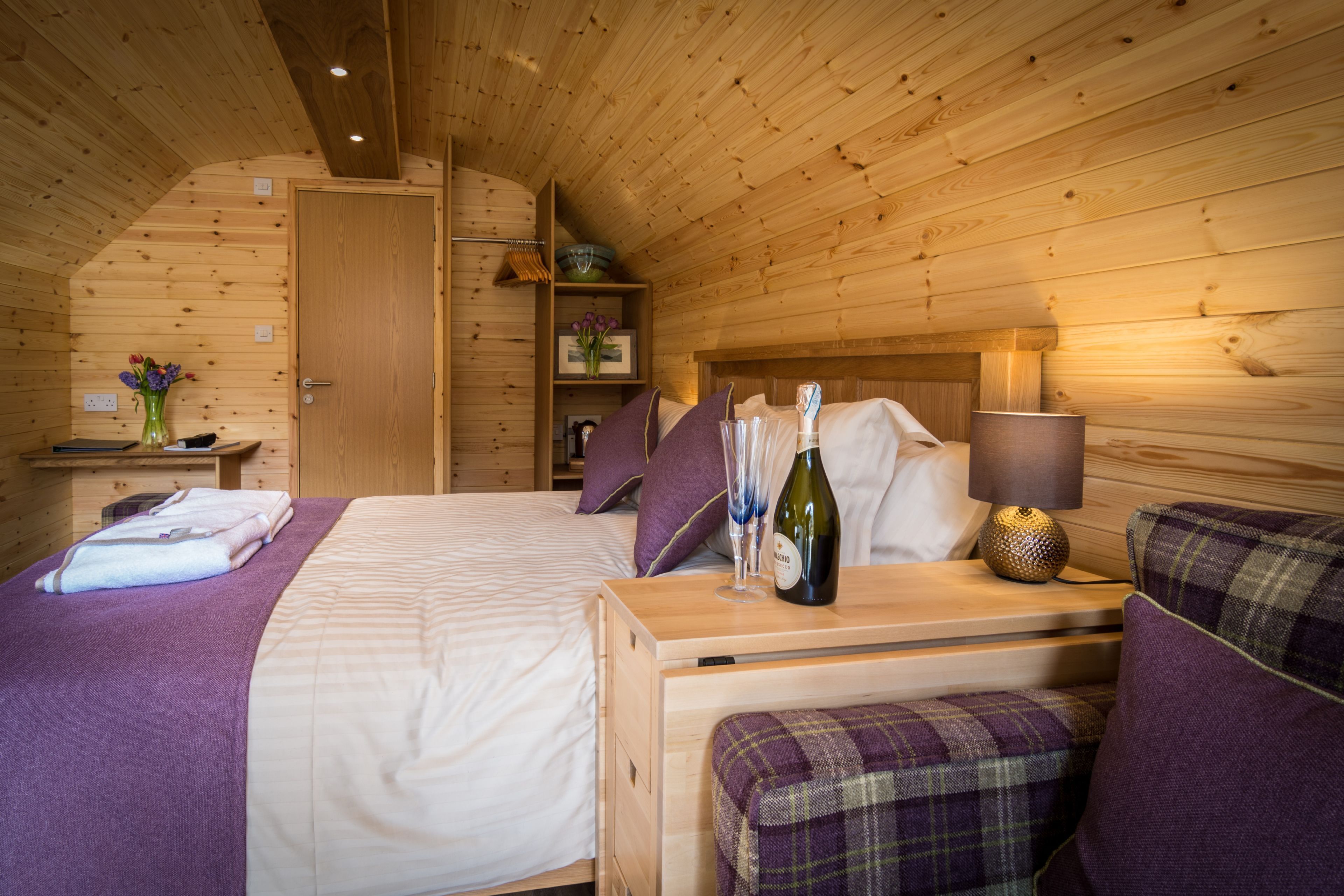 Un dormitorio de cabaña de madera con una cama, una mesita de noche con una botella y vasos, una lámpara y un banco con cojín.