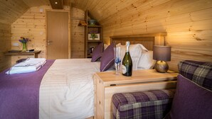 A wooden cabin bedroom with a bed, a bedside table with a bottle and glasses, a lamp, and a bench with a cushion. - Cosy Eco Cabin with stunning views (Glencoe)