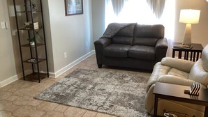 A living room with a dark leather sofa, a beige armchair, a small side table with a lamp, a bookshelf, and a framed picture on the wall.