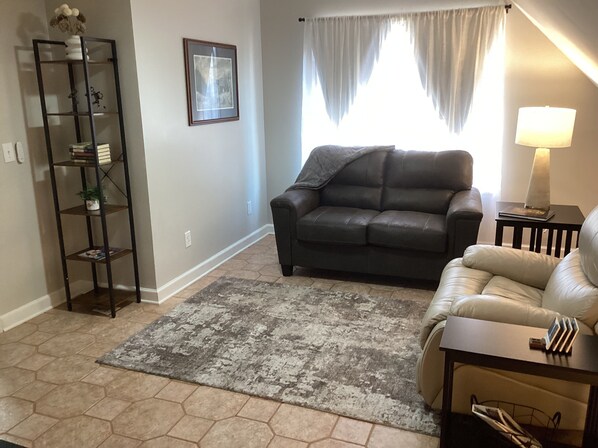 A living room with a dark leather sofa, a beige armchair, a small side table with a lamp, a bookshelf, and a framed picture on the wall.