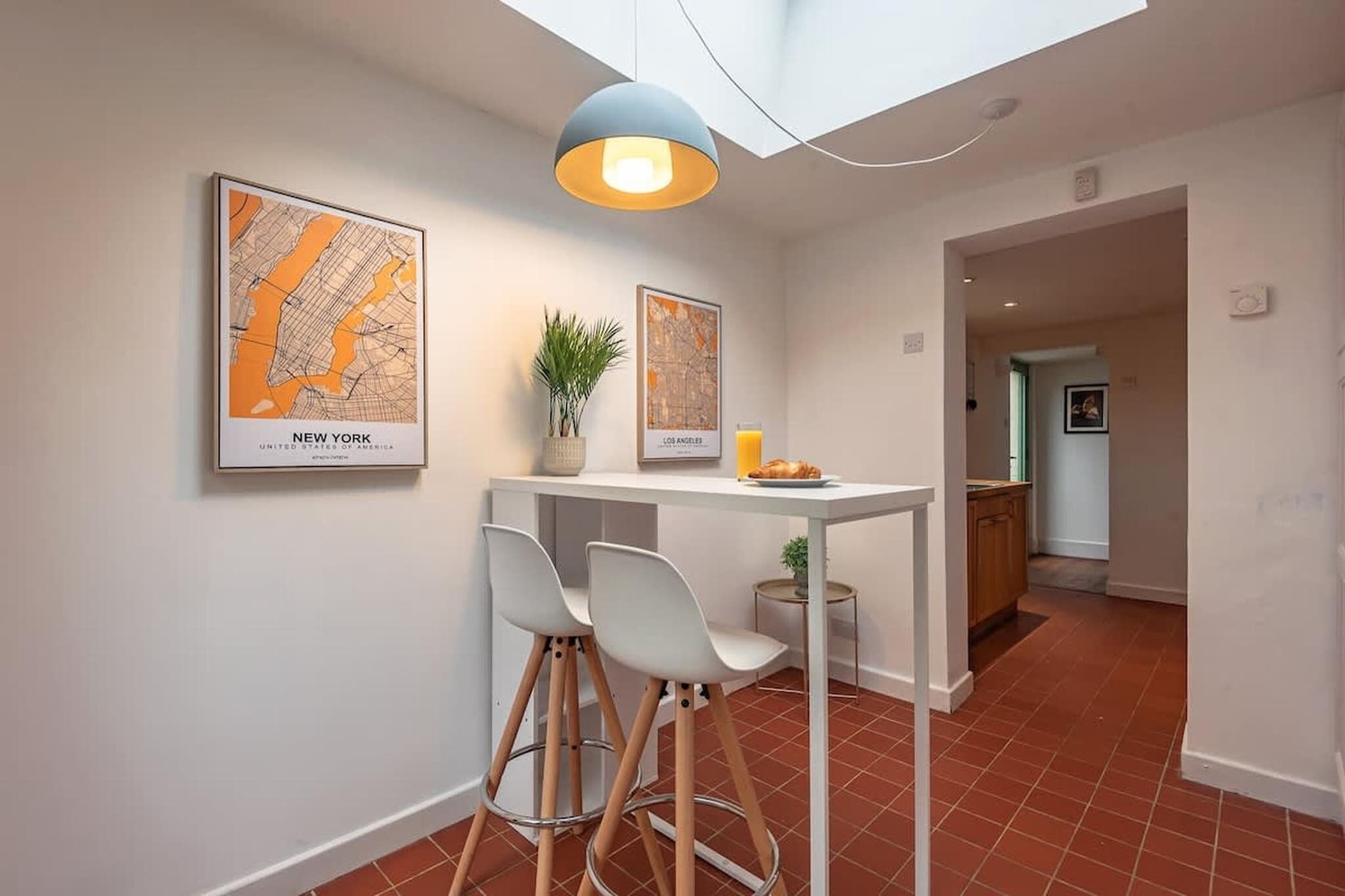Eine moderne Küche mit Essbereich, ausgestattet mit einem weißen Tisch, zwei Barhocker und einer Pendelleuchte. An der Wand hängen zwei gerahmte Karten, eine mit der Aufschrift „New York“ und die andere „Los Angeles“.