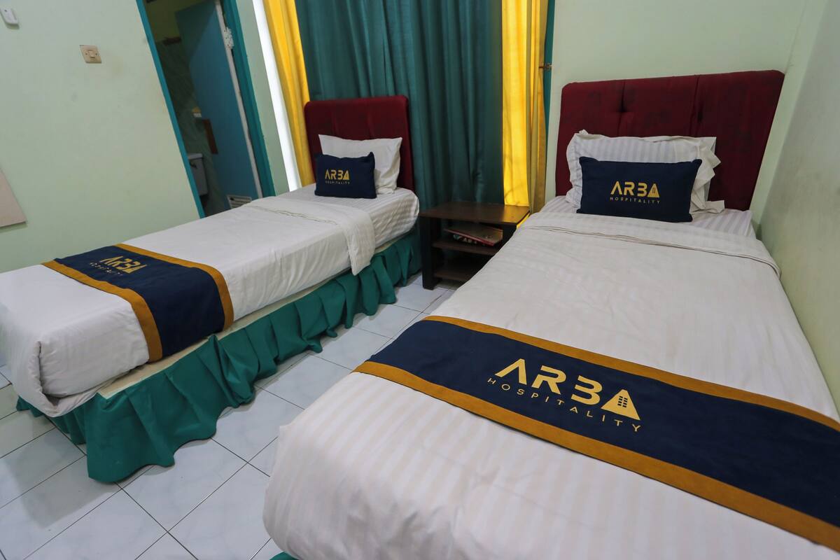 Dos camas individuales con cabeceras y pies de cama a juego, cada una con una almohada y una mesita de noche. Las camas presentan un diseño uniforme con un logotipo en la colcha.