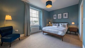 Ein Schlafzimmer mit einem Bett, einem Sessel, zwei Nachttischen mit Lampen, einem Fenster mit Vorhängen und gerahmten Bildern an der Wand.
