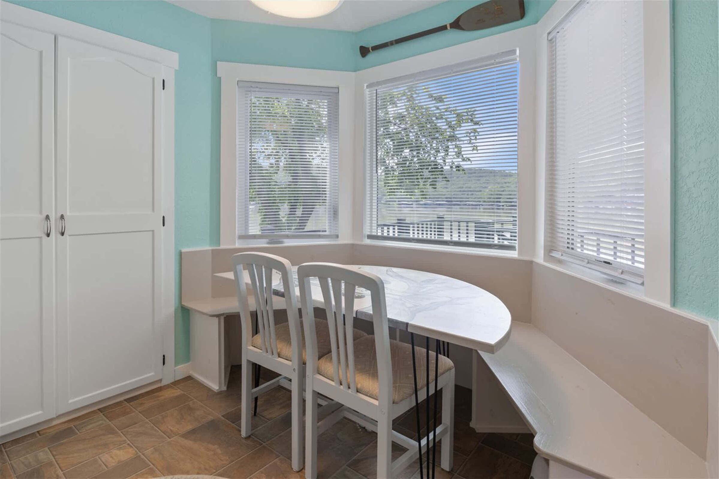 Een keuken met een witte tafel en twee stoelen, een raam met jaloezieën en een deur.