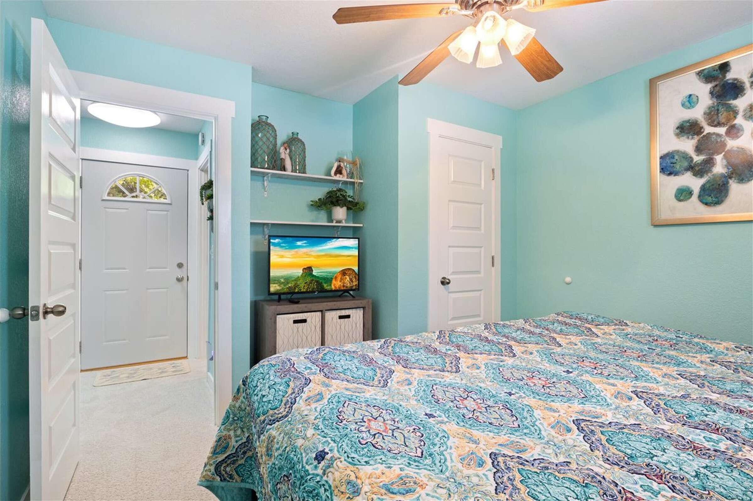 Un dormitorio con una cama, un ventilador de techo, un televisor sobre un mueble y una puerta con ventanal.