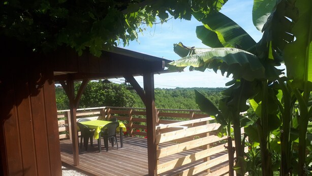 Une terrasse en bois avec une table et des chaises, entourée d’une végétation luxuriante et sous un ciel dégagé.