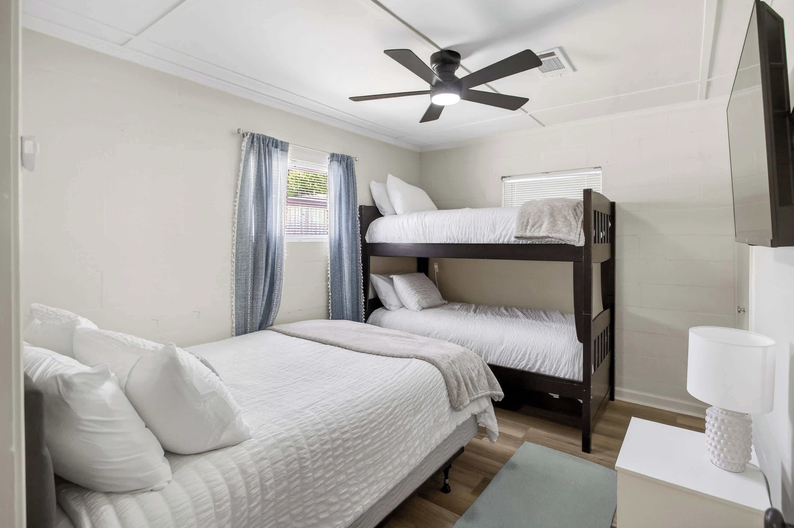 Une chambre avec deux lits simples, un lit superposé, un ventilateur de plafond, une fenêtre avec des rideaux et une lampe blanche sur une table de chevet.