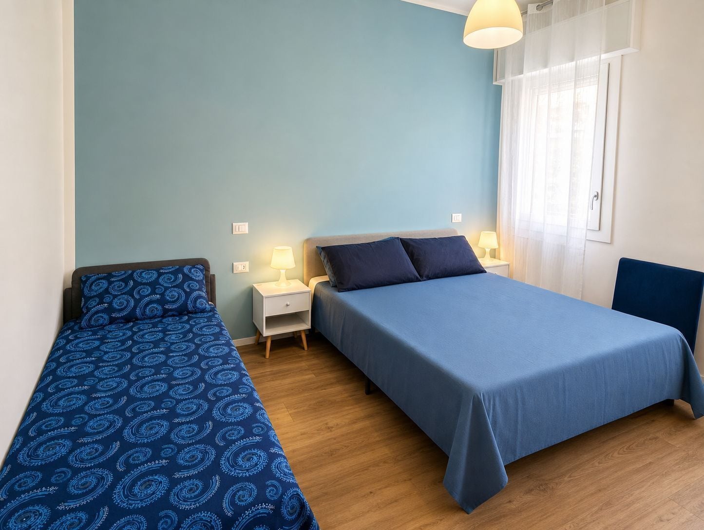 A bedroom with two beds, a nightstand, a lamp, and a window with curtains.