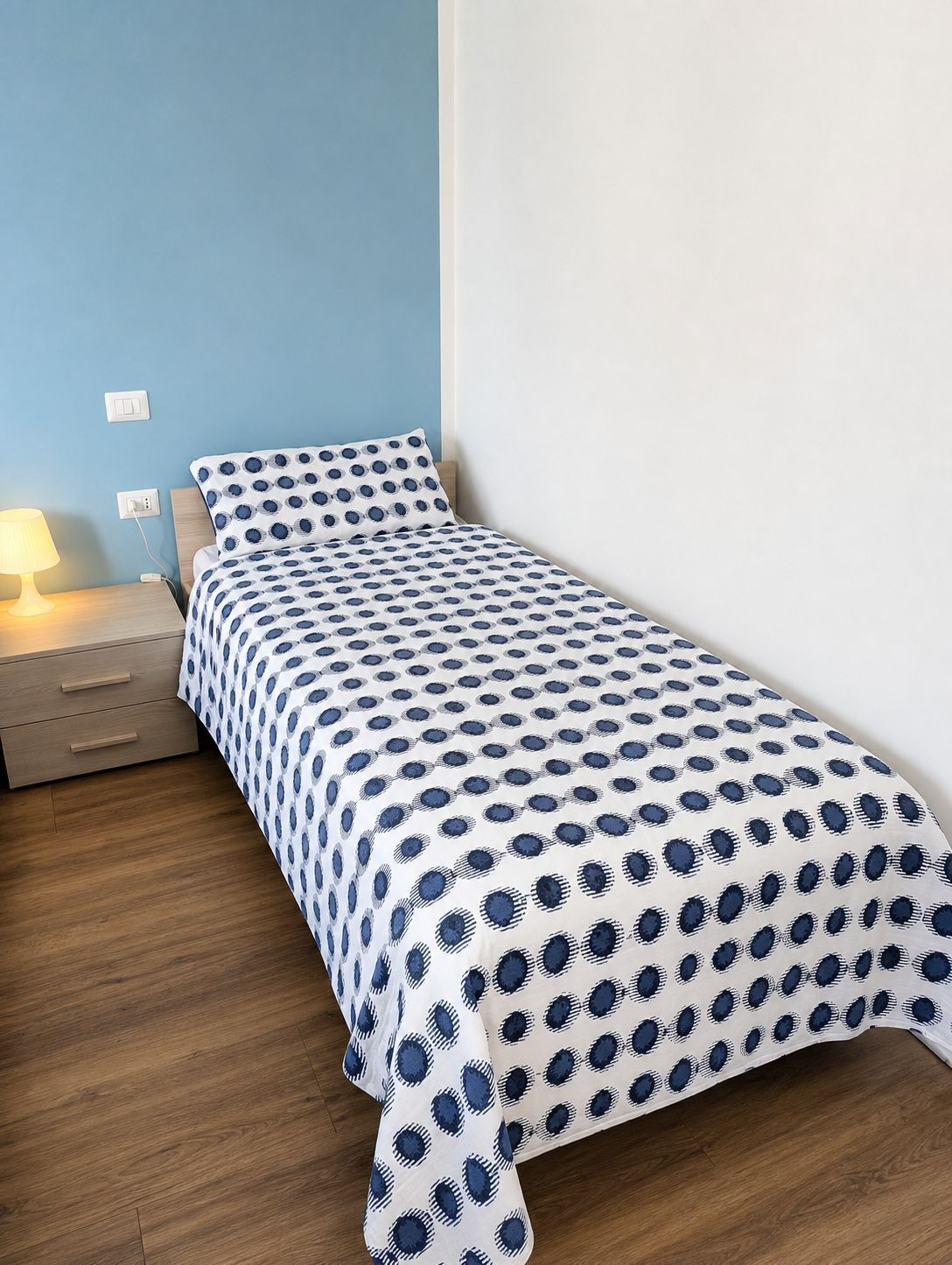 Una cama individual con una colcha estampada, una mesita de noche con lámpara y un enchufe en la pared.