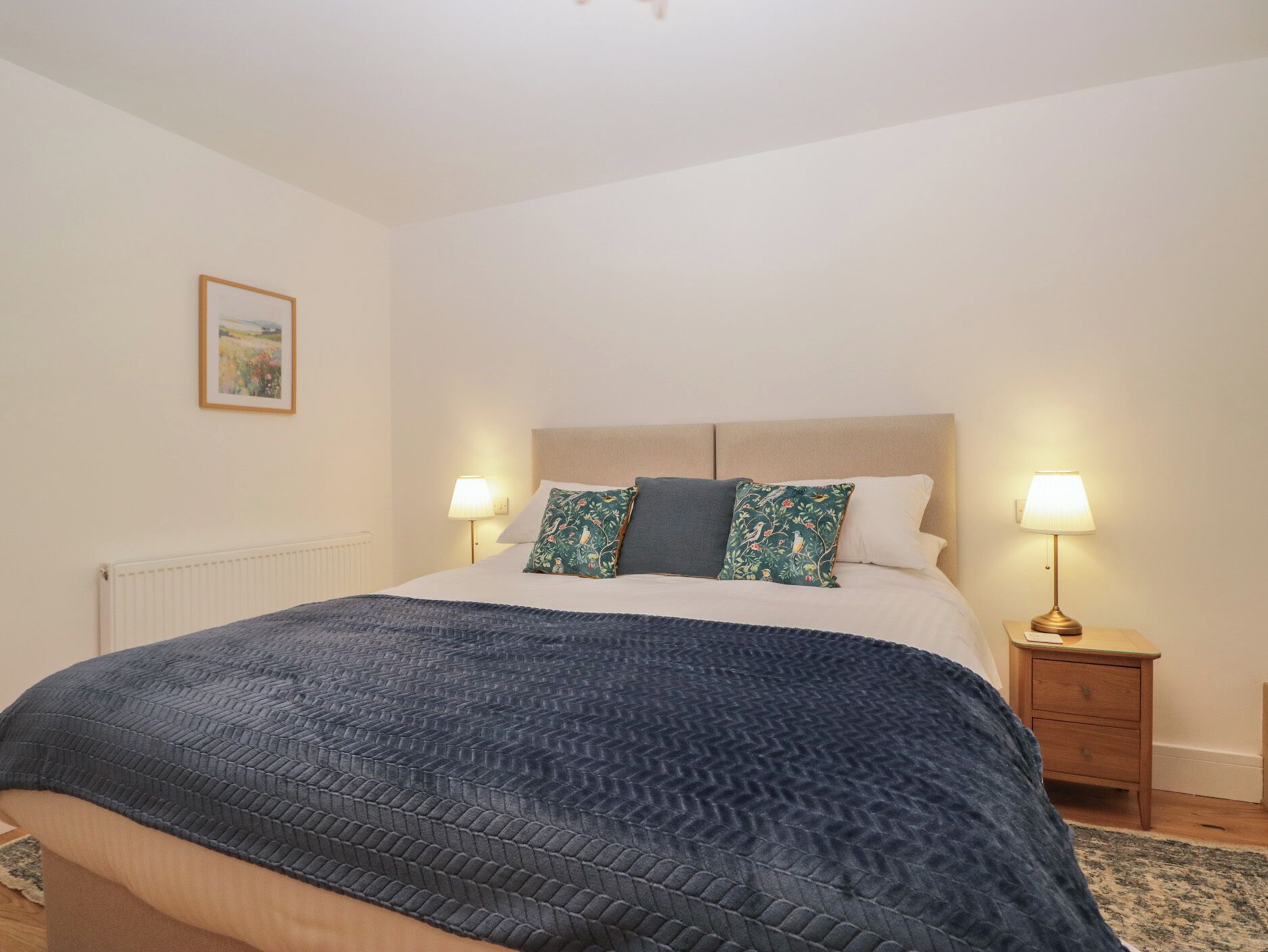A bedroom with a large bed, two bedside tables with lamps, a radiator, and a framed picture on the wall.