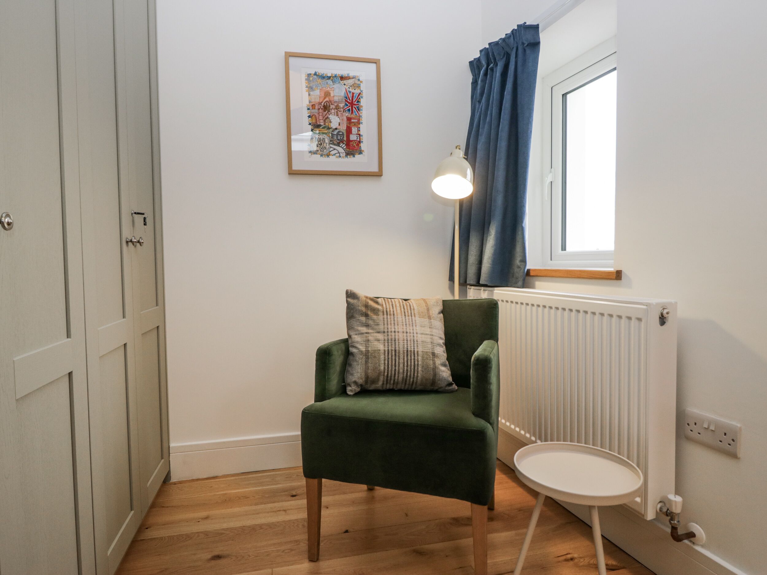 A room with a green chair, a small round table, a framed picture on the wall, a window with blue curtains, and a radiator.