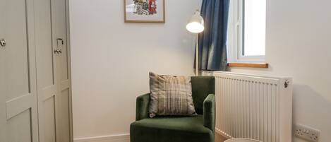 A room with a green chair, a small round table, a framed picture on the wall, a window with blue curtains, and a radiator.