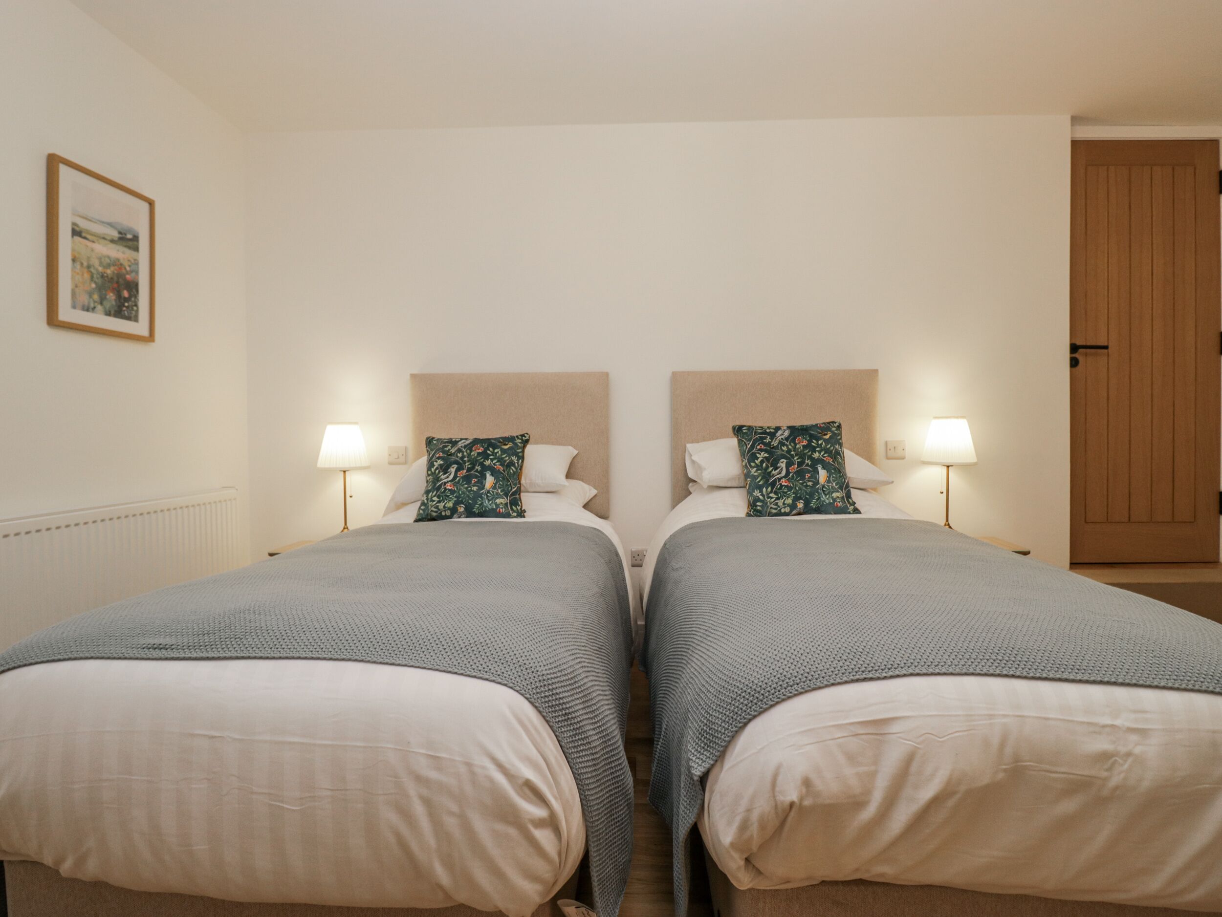 A double room with two beds, each with a headboard, a bedside table, a lamp, and a pillow.