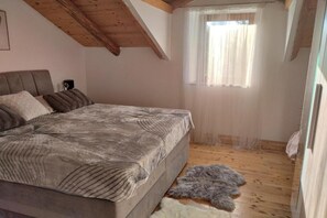 A bedroom with a bed, wooden ceiling beams, a window with sheer curtains, and a fur rug on the floor.