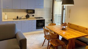 A compact kitchen with blue cabinets, a wooden dining table, and a sofa. - Natalia Cottage (Pieve di Ledro)