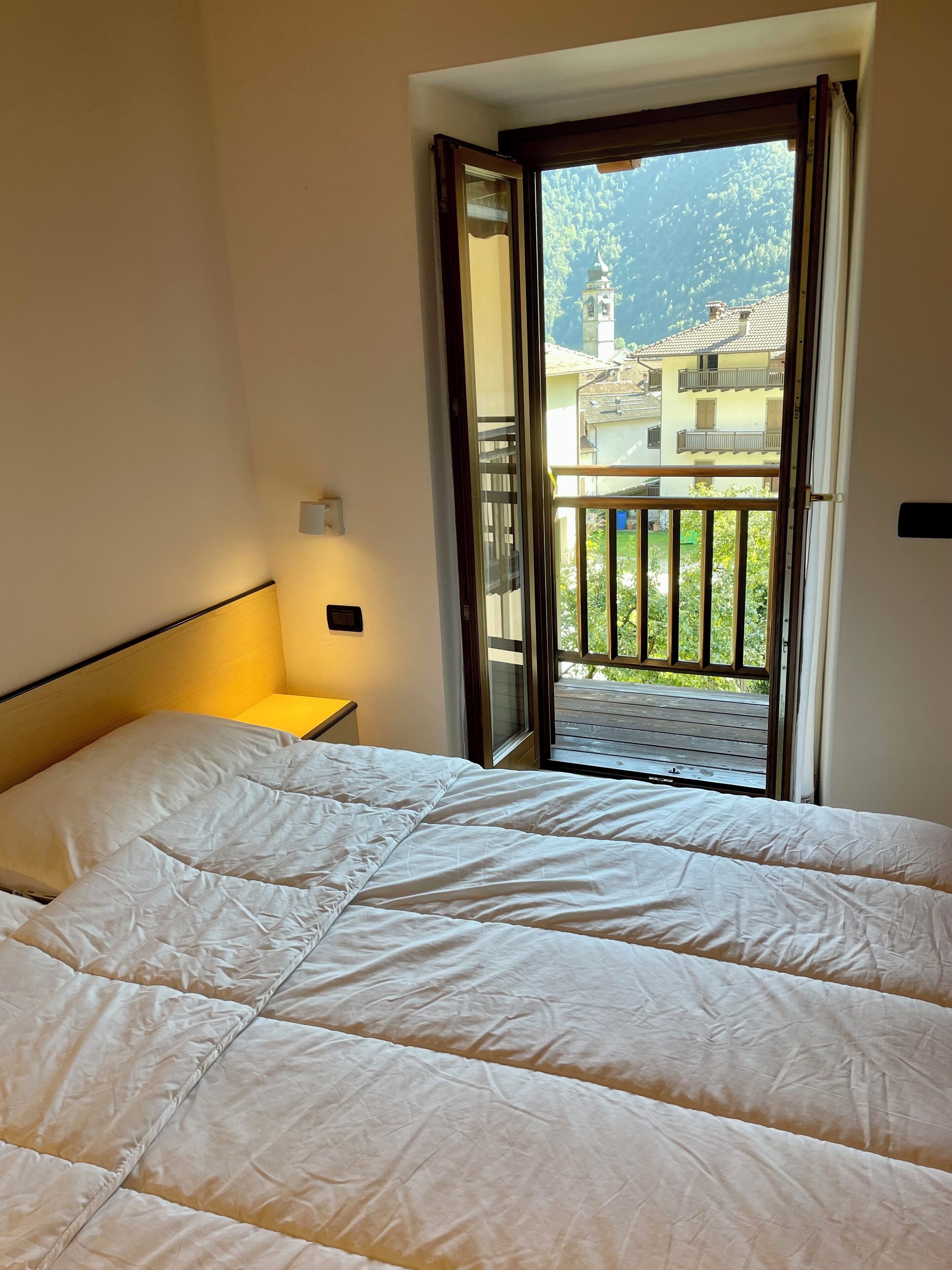 Ein Hotelzimmer mit einem großen Bett, einem Nachttisch und Blick auf einen Balkon mit Geländer sowie Gebäude im Hintergrund.