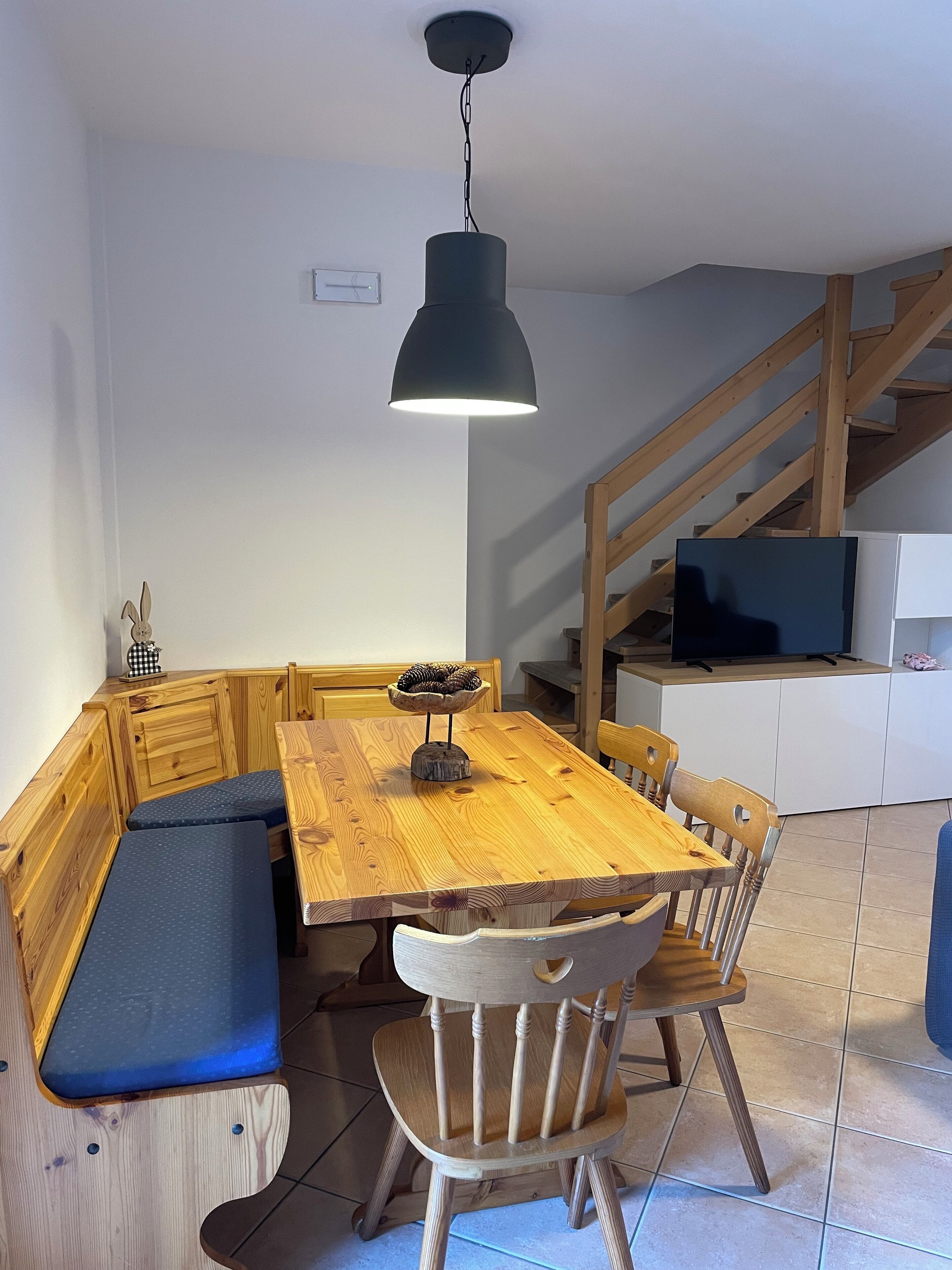 Un tavolo da pranzo in legno con panche, una lampada a sospensione nera e un televisore su un mobile.