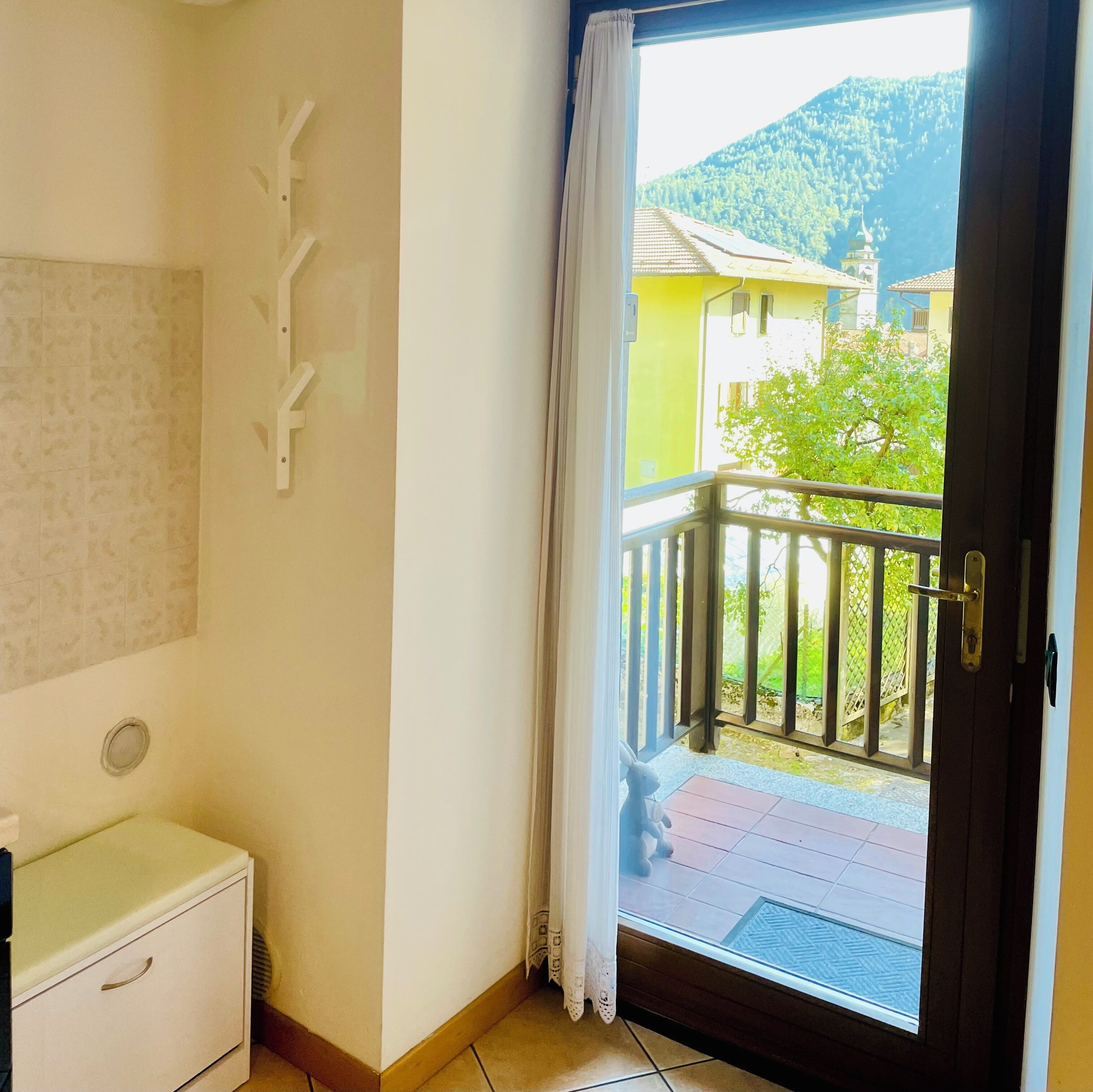 Ein Zimmer mit einem kleinen weißen Schrank, einer Wandleuchte und einer Glastür zum Balkon mit Blick auf einen Berg und ein Gebäude.