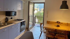 A compact kitchen with modern appliances, a dining area with a wooden table and chairs, and a balcony with a view of buildings and trees. - Natalia Cottage (Pieve di Ledro)