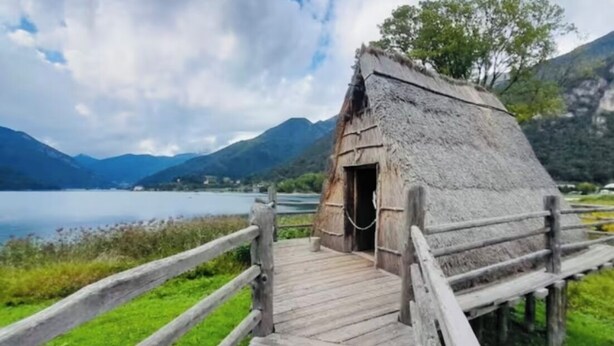 A wooden structure with a thatched roof, a wooden walkway leading to it, and a lake in the background. - Natalia Cottage (Pieve di Ledro)