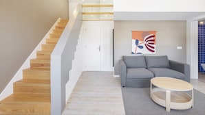 Un interior moderno con una escalera, un sofá gris, una mesa redonda de madera y un cuadro abstracto de colores.