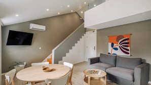 Un área de estar moderna con una mesa redonda de madera, sillas blancas, un sofá gris y un televisor de pantalla plana montado en la pared.