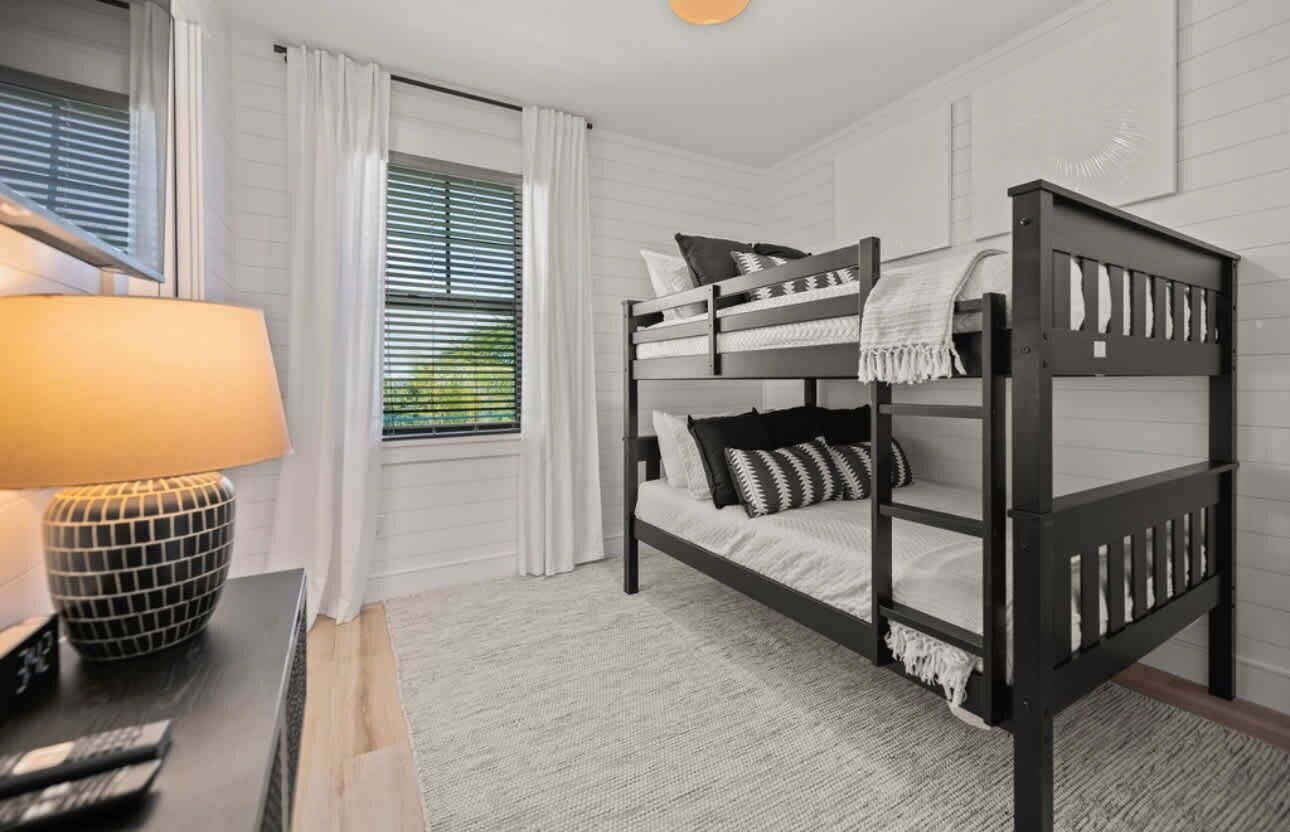 A bunk bed room with white curtains, a lamp, and a window with blinds.