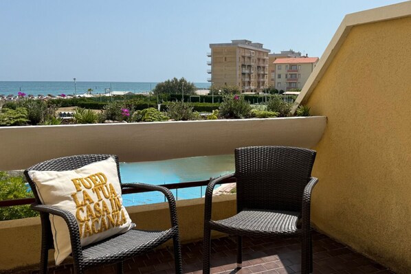 Ein Balkon mit zwei Korbstühlen, einem Pool und Blick auf das Meer und Gebäude.