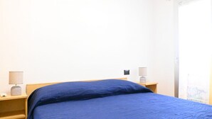 En enkeltseng med et blåt sengetæppe, to sengeborde med lamper, og en dør med udsigt over bybilledet.